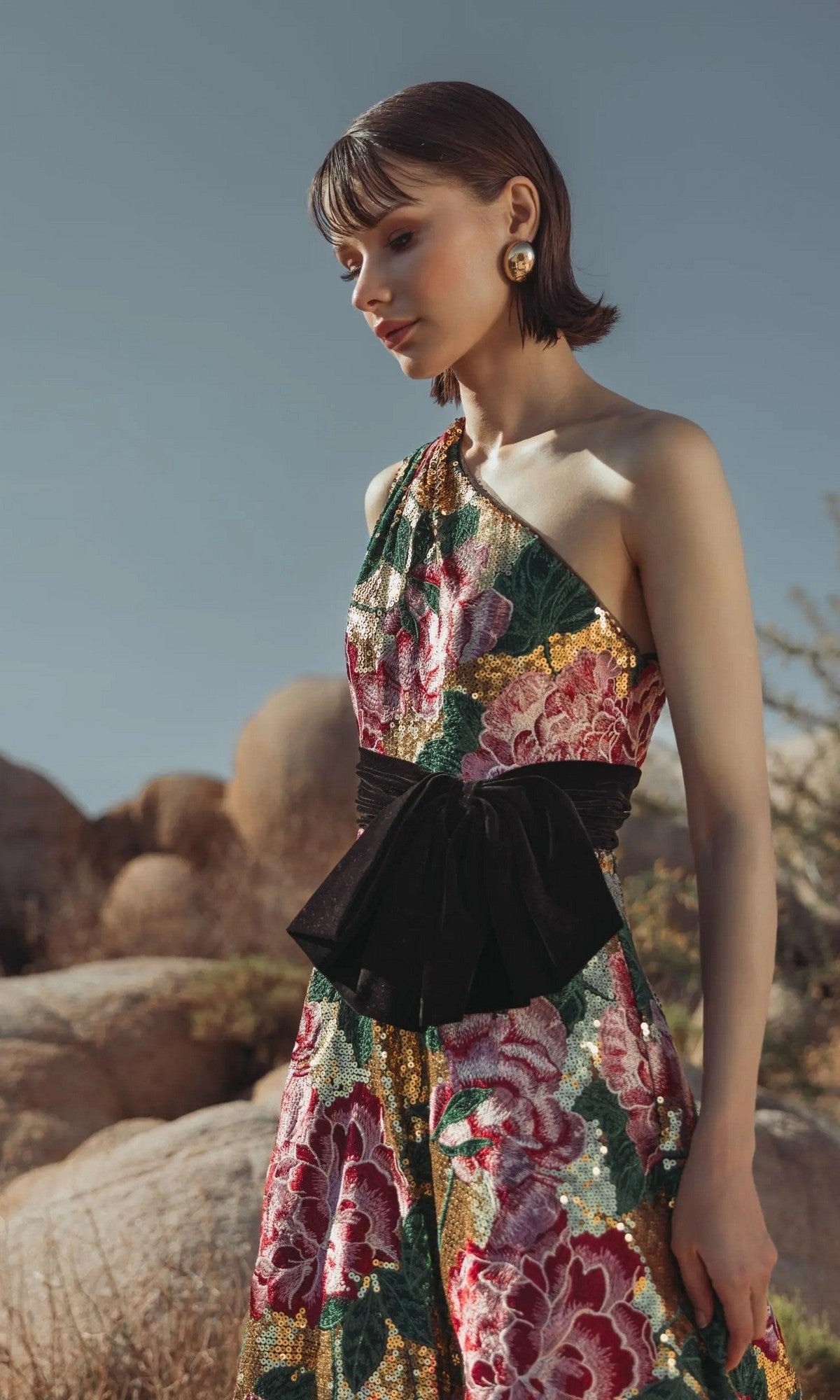 A woman stands outdoors near large rocks, looking down, wearing the Mac Duggal 21194 long formal dress featuring one shoulder, floral embroidery, and a black sash.